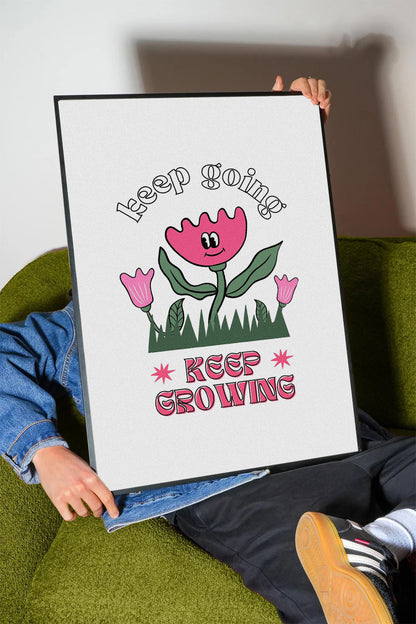Siyah çerçeveli, beyaz zeminde pembe gülen çiçek, küçük çiçekler ve çimenler ile 'Keep Going' ve 'Keep Growing' yazılı motivasyon posteri.