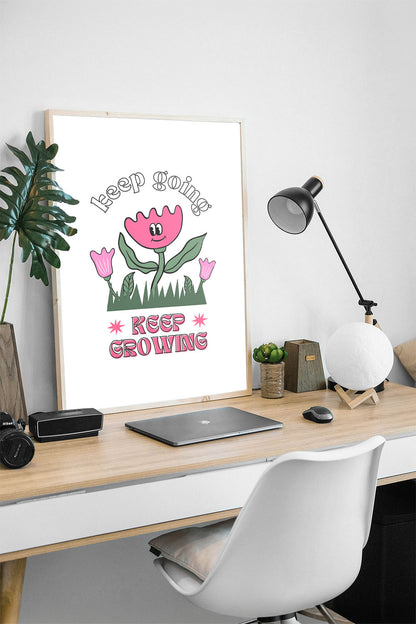 Siyah çerçeveli, beyaz zeminde pembe gülen çiçek, küçük çiçekler ve çimenler ile 'Keep Going' ve 'Keep Growing' yazılı motivasyon posteri.