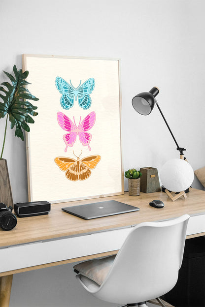 Siyah çerçeveli, açık renk dokulu zeminde mavi, pembe ve kahverengi üç modern kelebek desenli tablo.