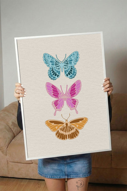Siyah çerçeveli, açık renk dokulu zeminde mavi, pembe ve kahverengi üç modern kelebek desenli tablo.