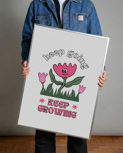 Siyah çerçeveli, beyaz zeminde pembe gülen çiçek, küçük çiçekler ve çimenler ile 'Keep Going' ve 'Keep Growing' yazılı motivasyon posteri.